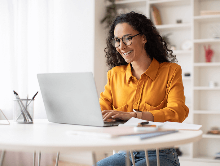 woman in mustard yellow shirt smiling while typing on laptop at home table.